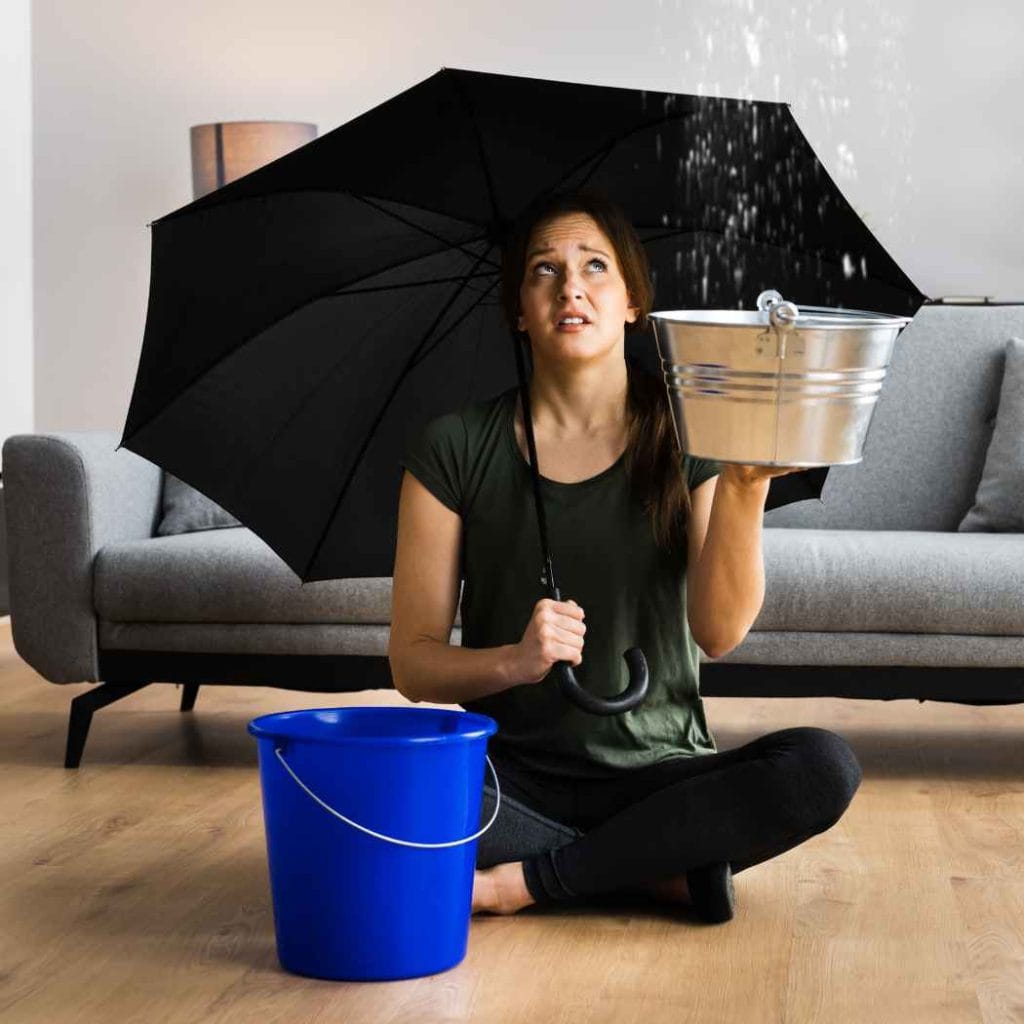 Frustrated woman sitting on floor holding an umbrella and a metal bucket to catch water leaking from ceiling during an indoor flood emergency.