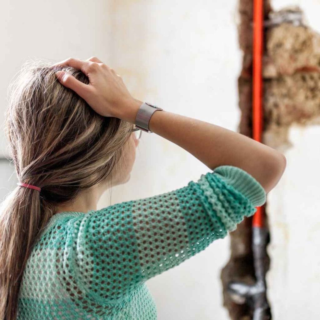 Worried woman looking at severe water damage and crumbling plaster on an interior wall caused by a burst pipe or flooding.