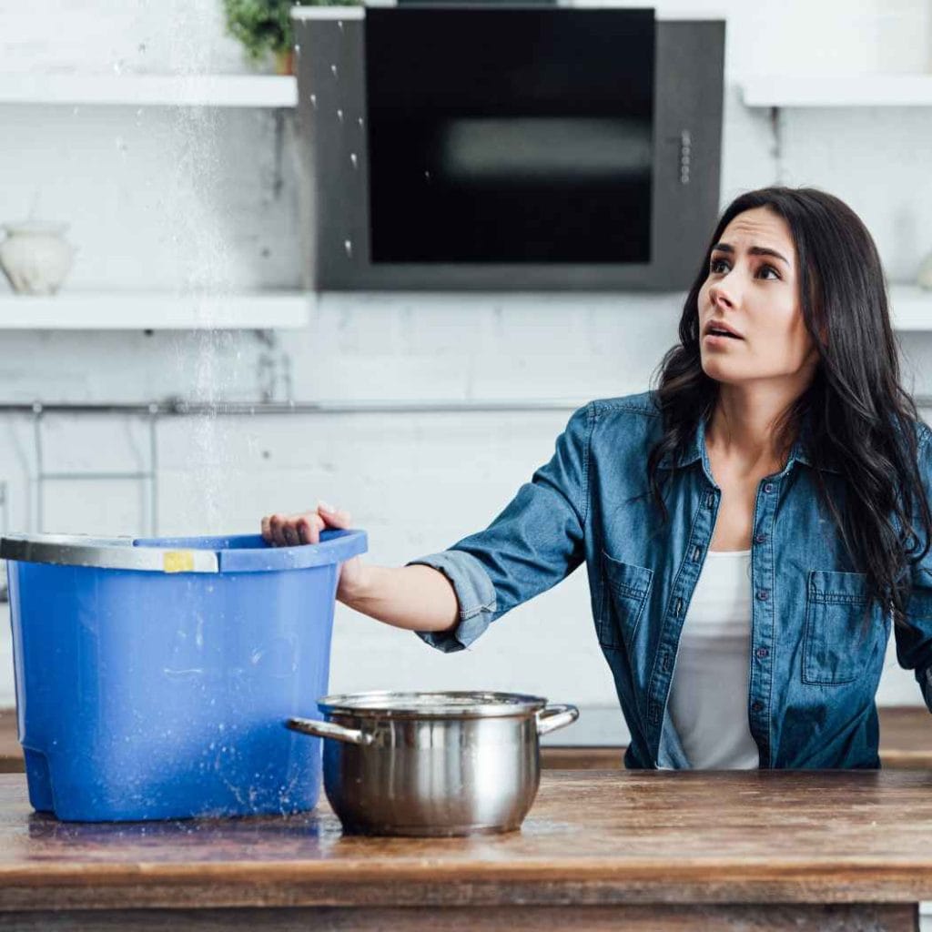Worried woman placing a bucket under a leaking ceiling to catch dripping water during an indoor water damage emergency.