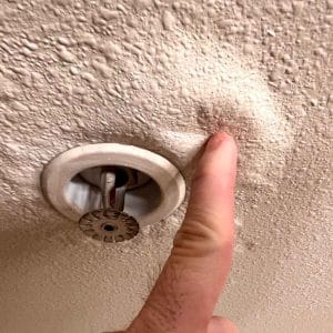 Close-up of a water-damaged ceiling with bubbling and sagging texture near a fire sprinkler head, as a finger presses against the swollen area indicating trapped moisture.