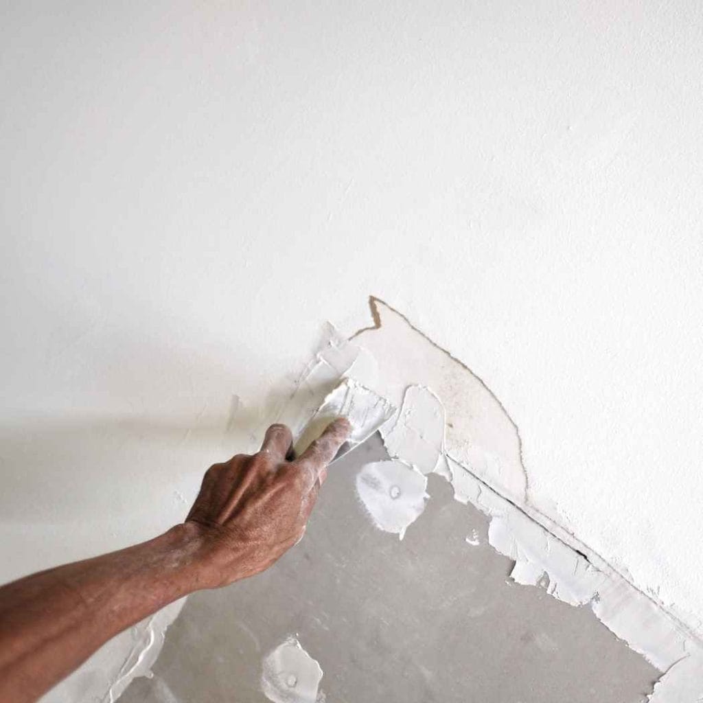 Close-up of a person scraping off damaged drywall caused by water infiltration, exposing underlying moisture-affected layers.