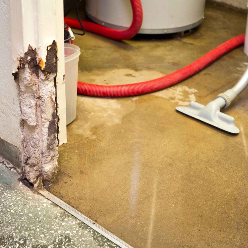 Flood-damaged laundry area with rusted door frame, wet concrete floor, red water extraction hose, and vacuum equipment used for water removal.