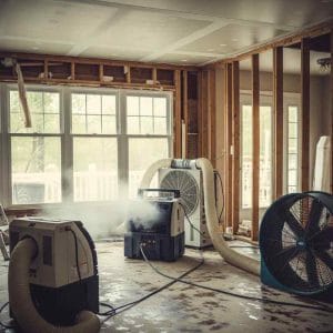Flood-damaged room with exposed wall framing and industrial drying equipment in operation, including air movers and dehumidifiers actively removing moisture from the stripped interior.
