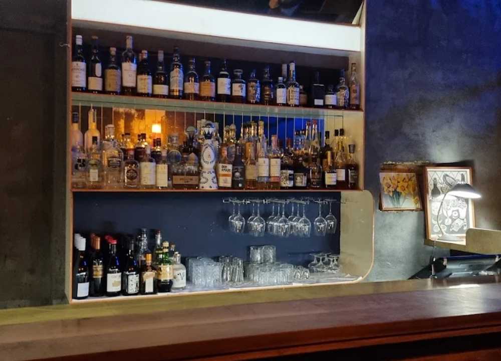 A stylish and intimate view of the bar at Angel Music Bar in Melbourne, showcasing neatly arranged shelves filled with premium spirits, wine bottles, and hanging glassware. The ambient lighting casts a warm glow, while framed art and a desk lamp add a cosy, artistic touch to the modern, minimalist décor.