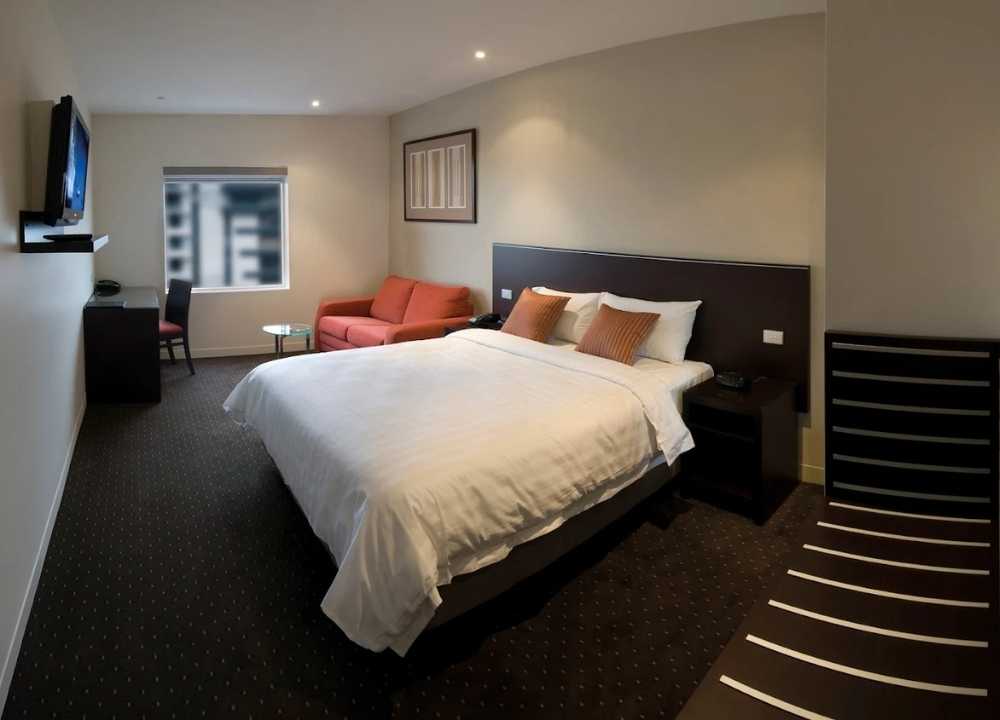 A comfortable and modern guest room at Atlantis Hotel in Melbourne, featuring a large king-size bed with white linens and orange accent pillows. The room includes a red sofa, a glass coffee table, a wall-mounted TV, a work desk, and a window with a city view. Neutral tones and simple furnishings create a welcoming and practical space for travellers.