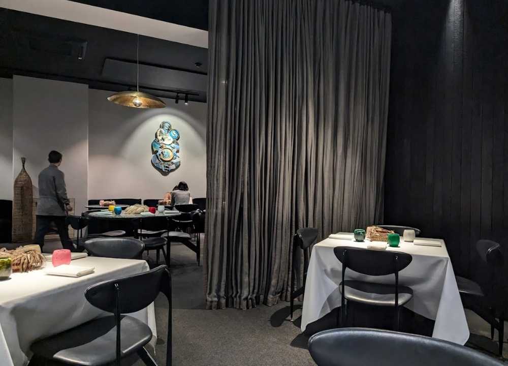 A sophisticated dining area at Attica restaurant in Melbourne, featuring neatly set tables with white cloths, modern black chairs, and soft ambient lighting. A large curtain divides the space, and contemporary wall art adds character to the minimalist interior. Diners are seen enjoying their meals in the background.