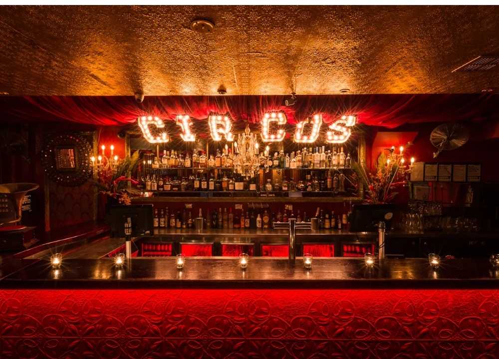 Interior of Circus Bar, a stylish and theatrical nightclub in Melbourne, featuring a dimly lit bar with ornate red decor and vintage flair. The word "CIRCUS" is illuminated in marquee-style lights above shelves stocked with bottles of liquor. Chandeliers, candlelight, and velvet curtains enhance the venue’s rich, cabaret-inspired atmosphere.