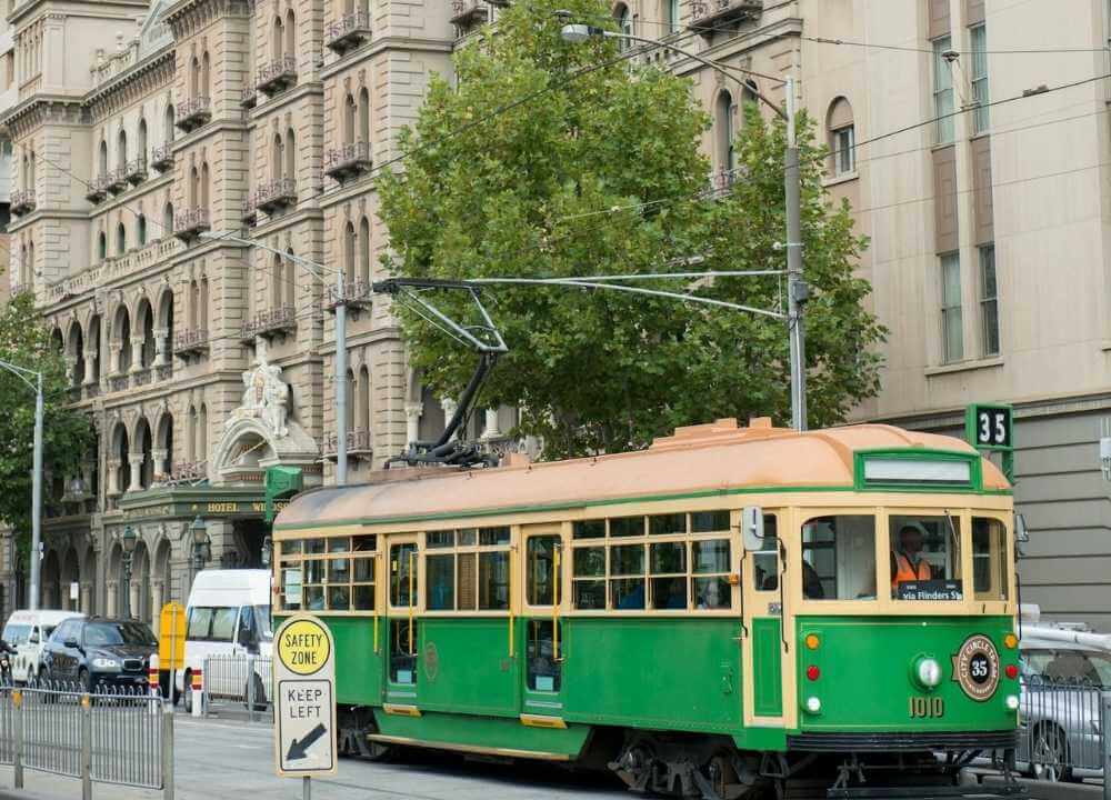 A historic green and cream City Circle Tram (route 35) travels past the ornate Hotel Windsor building in Melbourne. The vintage tram is a free tourist service, and the scene captures a blend of classic architecture, urban street life, and greenery from nearby trees. A sign in the foreground reads "Safety Zone – Keep Left.