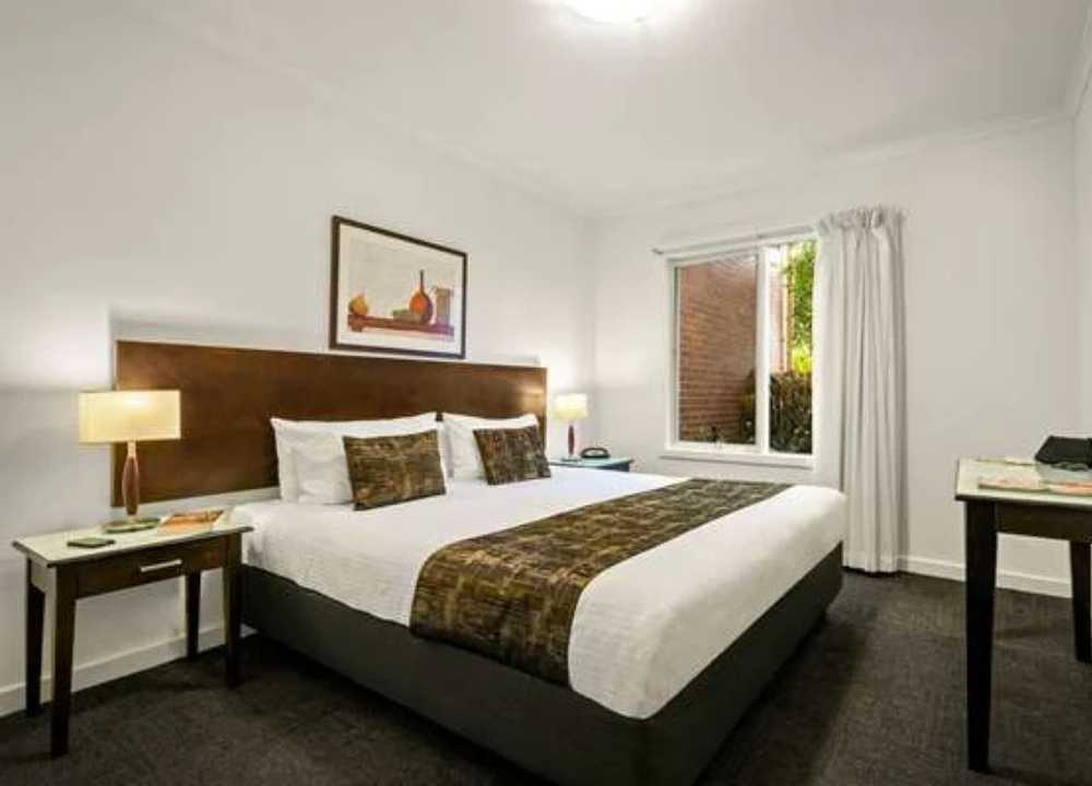 A clean and cosy bedroom at Comfort Apartments Royal Gardens in Melbourne, featuring a king-sized bed with white linens, decorative brown cushions and runner, modern bedside lamps, a framed artwork above the headboard, and a window with white curtains letting in natural light. The room also includes a dark wooden desk and neutral-toned carpeting.