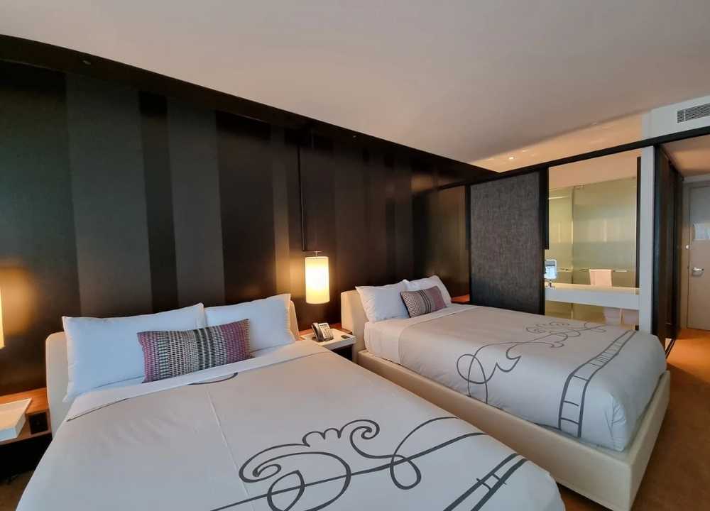 A stylish and modern twin room at Crown Metropol Melbourne, featuring two queen-sized beds with crisp white linens and elegant grey decorative patterns. The room has a sleek black accent wall, pendant bedside lights, a wooden floor, and a frosted-glass bathroom wall that adds a touch of luxury and openness to the contemporary design.