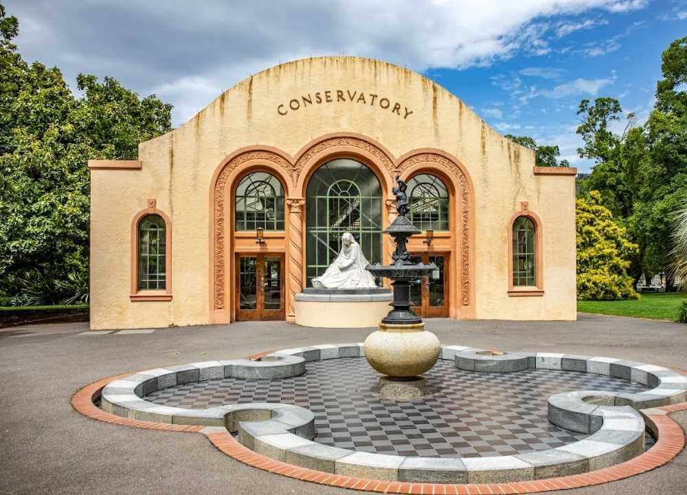 Front view of the Conservatory in Fitzroy Gardens, Melbourne, showcasing its elegant cream and terracotta Spanish Mission-style architecture. A statue of a seated woman is displayed at the entrance, framed by arched windows. In the foreground, a decorative fountain with a checkerboard-tiled base adds charm to the tranquil garden setting surrounded by lush greenery.