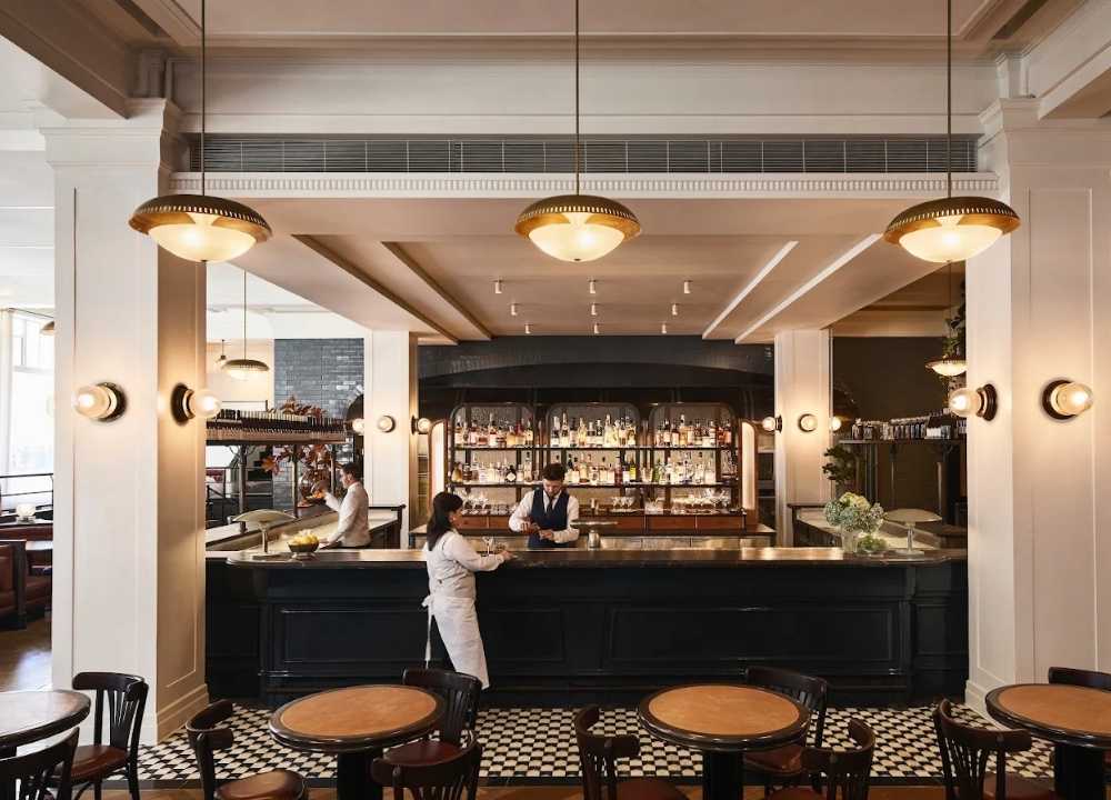 Chic interior of Gimlet at Cavendish House in Melbourne, featuring a stylish central bar with vintage lighting, black-and-white tiled flooring, round wooden tables, and classic decor. Bartenders and servers are seen preparing drinks in an elegant, upscale setting.
