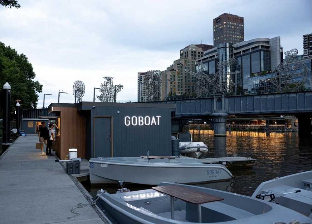 A twilight view of the GoBoat dock along the Yarra River in Melbourne, featuring a modern floating office with "GOBOAT" illuminated on its exterior. Several electric rental boats are moored nearby, ready for hire. In the background, city buildings and a decorated bridge with wire sculptures reflect in the calm water, creating a scenic urban riverside atmosphere.