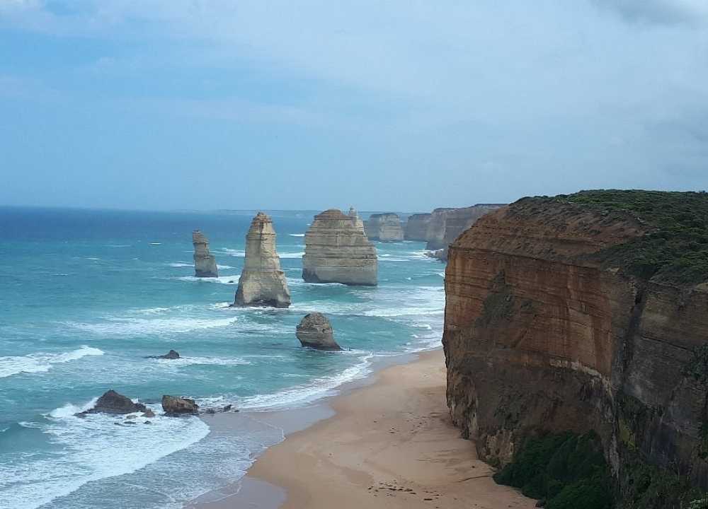 A stunning coastal view along the Great Ocean Road in Victoria, Australia, showcasing the iconic limestone stacks known as the Twelve Apostles rising dramatically from the turquoise waters of the Southern Ocean. Sheer cliffs frame the sandy shoreline, with waves gently crashing against the rock formations under a cloudy sky.