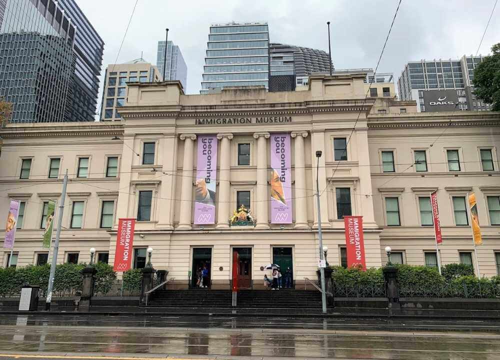 Front view of the Immigration Museum in Melbourne, housed in a grand neoclassical building with cream-coloured walls and tall columns. Large banners promoting the "Becoming You" exhibition hang between the columns, and red signs flank the entrance. The street and pavement are wet from recent rain, and modern skyscrapers tower in the background, blending history with urban life.