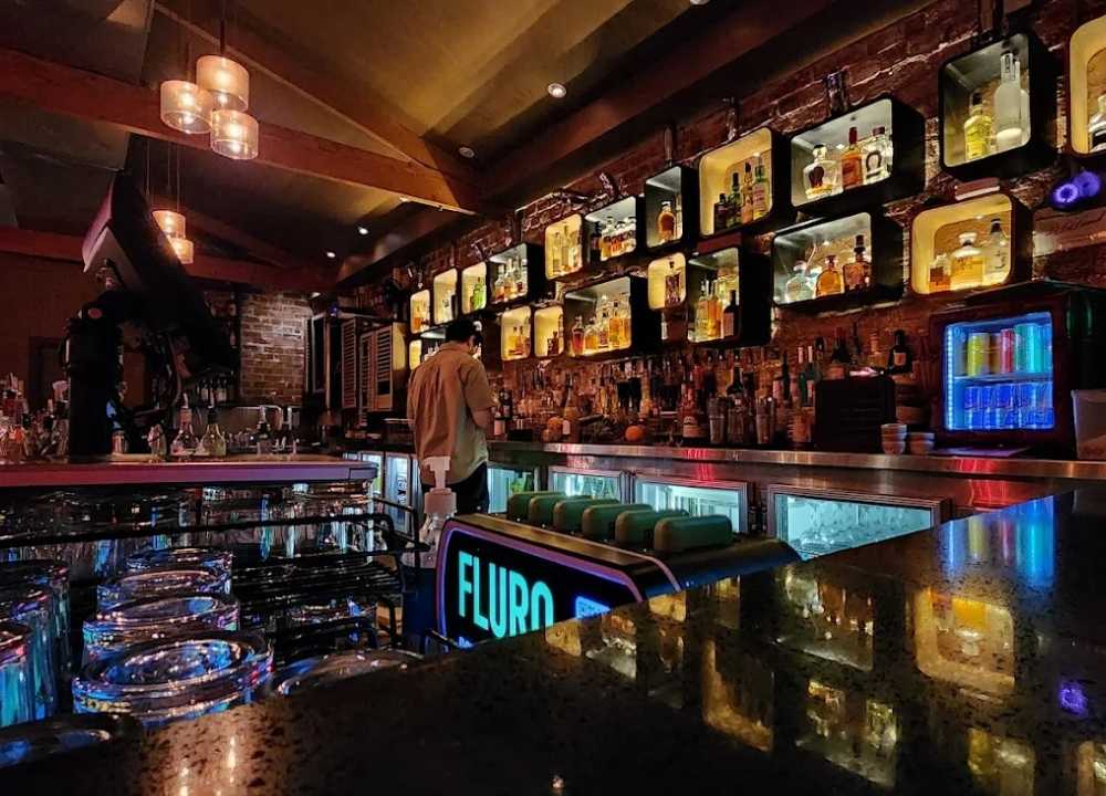 Interior of La La Land, a trendy bar and nightclub in Melbourne, featuring a sleek, modern bar counter with a glowing “FLURO” sign and rows of clean glassware. The back wall showcases an eclectic display of liquor bottles in illuminated cubby shelves against exposed brick. Warm lighting and a relaxed atmosphere create a stylish setting for late-night drinks.