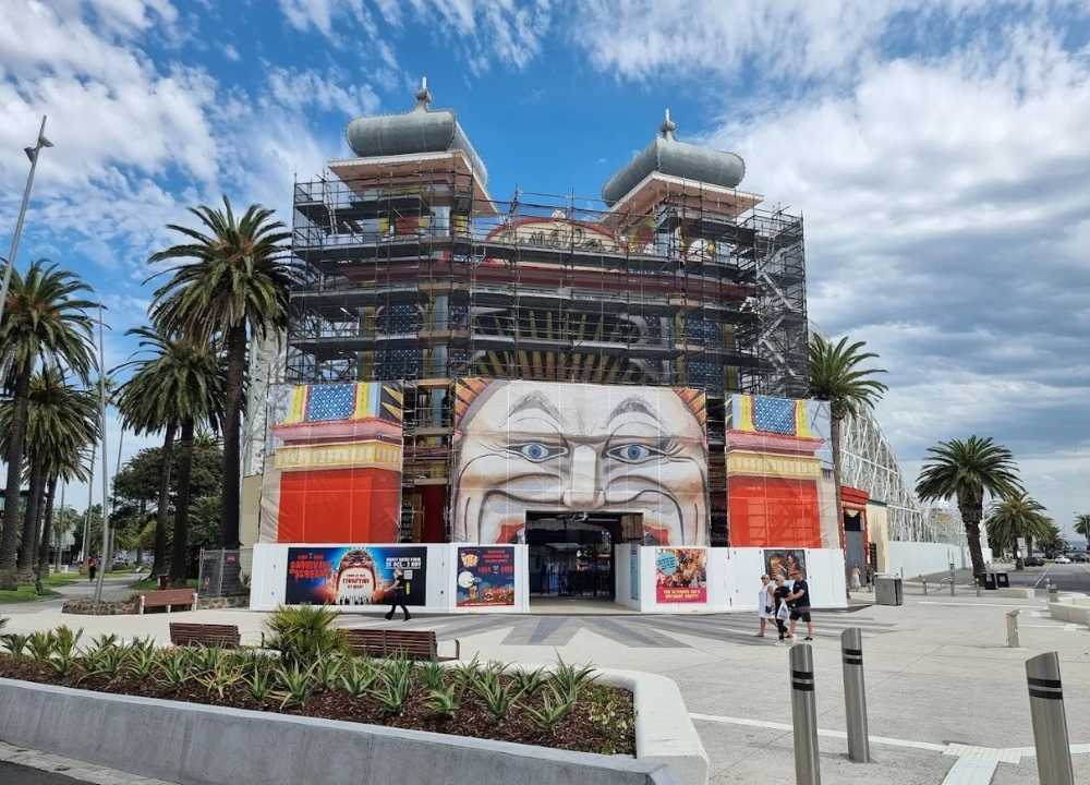 Front view of Luna Park in St Kilda, Melbourne, with its iconic giant smiling face entrance partially covered by scaffolding for renovation. Colorful banners and posters promote upcoming attractions, while palm trees line the walkway and a few visitors stroll past. The bright sky and coastal setting reflect the park’s classic seaside amusement charm.
