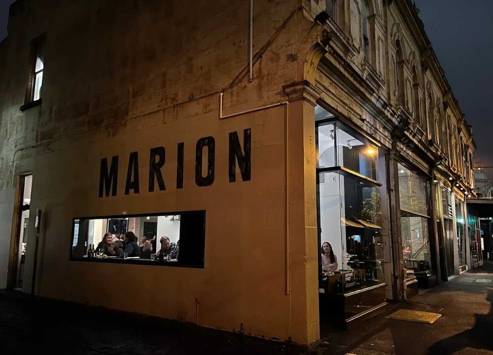 Nighttime exterior of Marion Wine Bar in Melbourne, showcasing its rustic, heritage-style building with bold black "MARION" lettering on the wall. Warm lighting spills through large windows, revealing diners inside enjoying food and wine in a relaxed, intimate atmosphere.