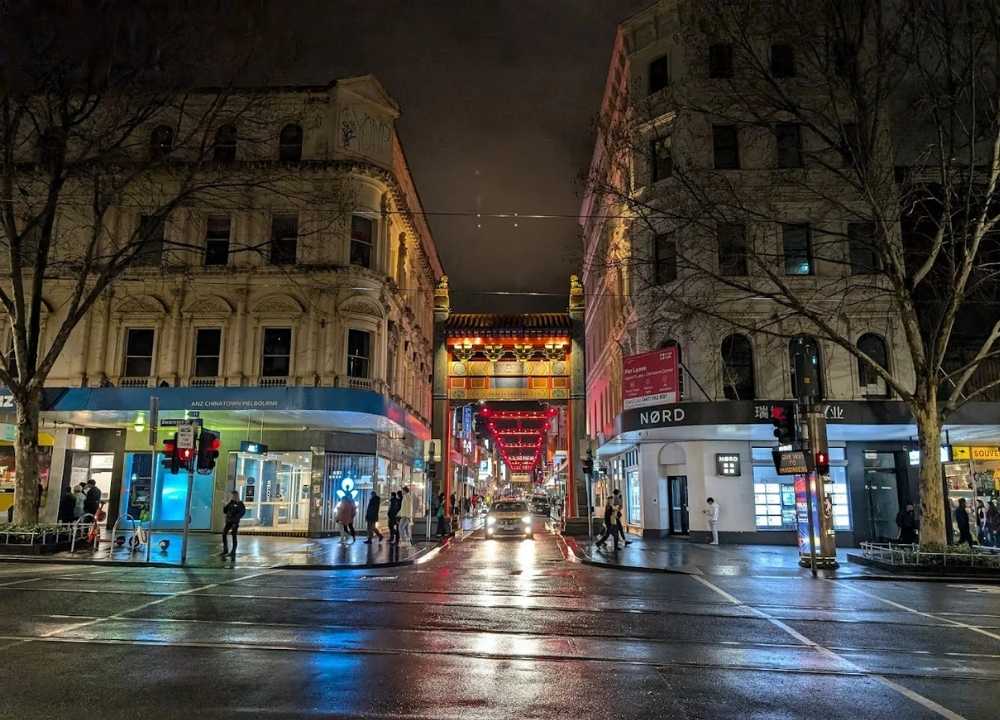 A vibrant night scene of Chinatown in Melbourne, featuring a traditional Chinese archway illuminated with red lanterns that stretches across the street. The wet pavement reflects the city lights, while pedestrians walk along the sidewalks and wait at the intersection. Historic buildings and modern shops, including ANZ and NØRD, line both sides of the street, adding to the lively urban atmosphere.
