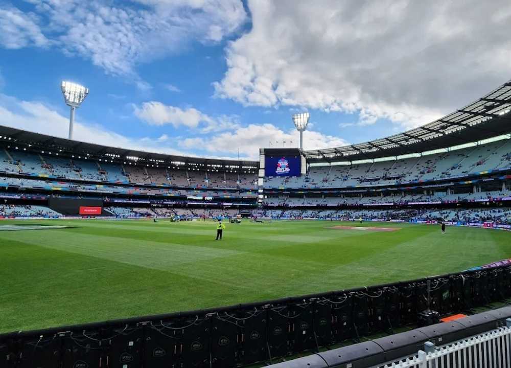 Wide-angle view of the Melbourne Cricket Ground (MCG) during a daytime event, showcasing its massive seating capacity and well-maintained green pitch. A few players and staff are seen on the field, with spectators scattered throughout the stands. Four tall floodlights tower over the stadium, and a large digital screen displays event graphics under a partly cloudy sky.