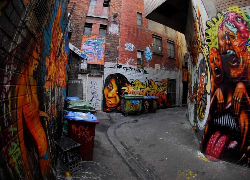 A colourful graffiti-filled laneway in Melbourne’s iconic arcades and laneways district. The narrow alley is adorned with bold street art on every brick wall, including vibrant portraits, abstract shapes, and surreal creatures. Several garbage bins line the path, adding an urban, gritty charm. The scene captures the raw creativity and cultural vibrancy of Melbourne's famous street art culture.