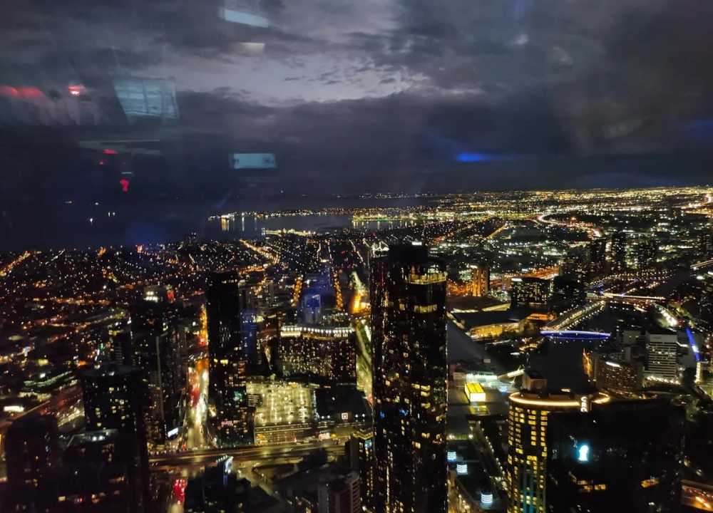 A breathtaking night view of Melbourne city from the Melbourne Skydeck, showcasing a sea of illuminated buildings, streets, and bridges stretching toward the horizon. The Yarra River winds through the glowing urban landscape, and Port Phillip Bay is visible in the distance under a dark, cloud-filled sky. The photo is taken through glass, with faint interior reflections visible.