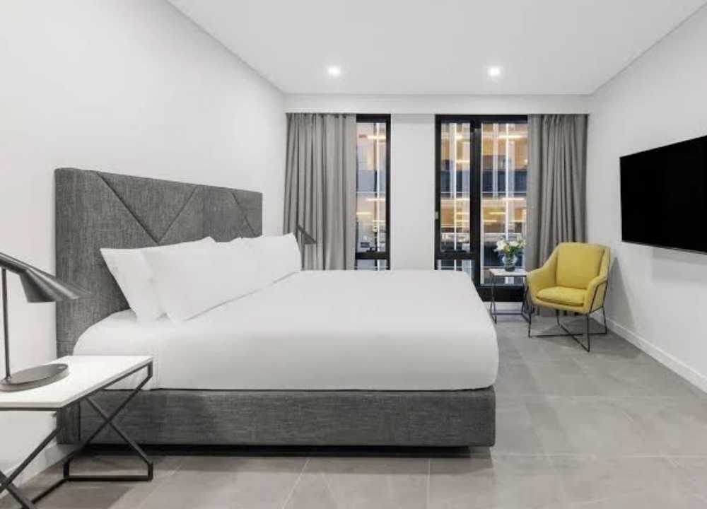 A sleek and spacious guest room at Meriton Suites in Melbourne, featuring a large grey-upholstered bed with crisp white bedding. The minimalist design includes a modern bedside table with a lamp, a yellow accent chair, a flat-screen TV mounted on the wall, and floor-to-ceiling windows with grey curtains, allowing natural light to fill the bright and contemporary space.