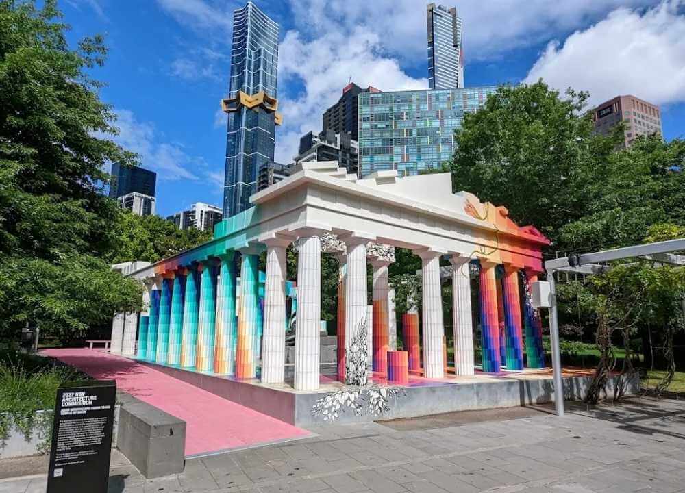 A colourful outdoor art installation near the National Gallery of Victoria in Melbourne, featuring a classical Greek-style structure with white columns transformed by vibrant rainbow gradients. The creative fusion of ancient architecture and modern art stands against a backdrop of tall skyscrapers and lush greenery under a bright blue sky with scattered clouds.