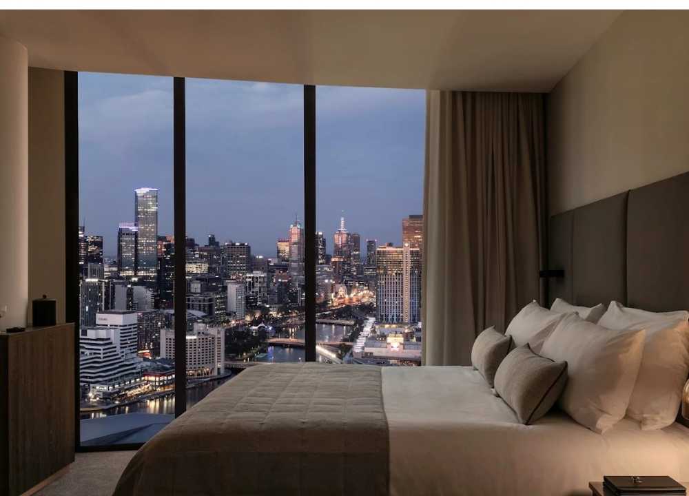 A luxurious bedroom at Oakwood Premier Melbourne, featuring a plush bed with neutral-toned bedding and pillows, set beside floor-to-ceiling windows offering a stunning panoramic view of Melbourne’s city skyline at dusk, with twinkling lights and high-rise buildings reflecting off the Yarra River.