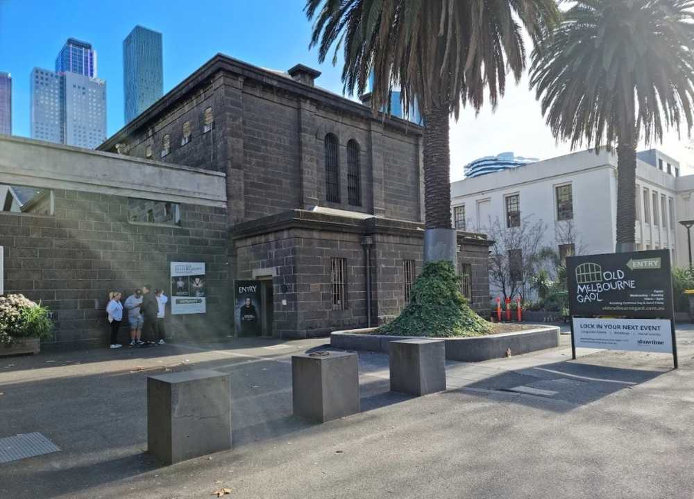 The exterior of Old Melbourne Gaol, a historic bluestone building under clear blue skies, framed by tall palm trees. A group of visitors stands near the entrance beneath a sign advertising entry to the museum. Modern city skyscrapers rise in the background, contrasting with the prison’s 19th-century architecture. A large sign on the right promotes the venue for events and tours.