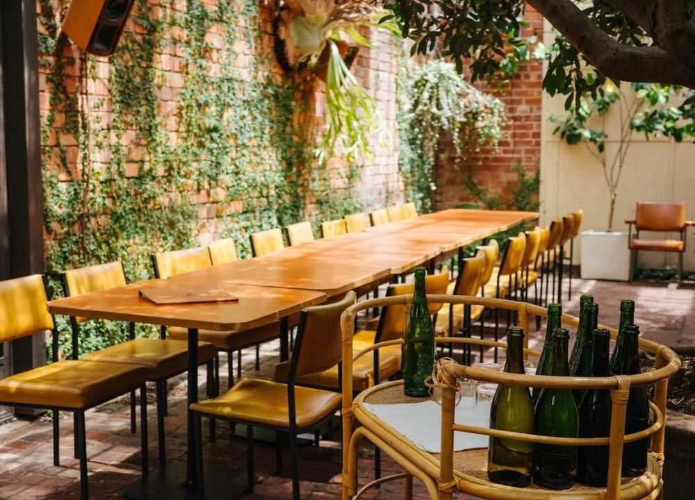 Outdoor dining area at Old Palm Liquor in Melbourne, featuring a long wooden communal table surrounded by mustard-yellow chairs. The space is adorned with lush green vines climbing up a rustic red-brick wall, creating a warm and inviting atmosphere. A wicker drinks trolley with empty green wine bottles adds to the relaxed, retro-inspired aesthetic.