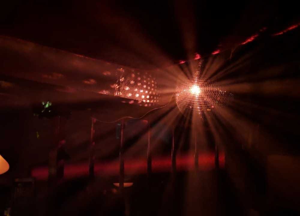 A glowing disco ball at Club OneSixOne in Melbourne emits dazzling beams of red and gold light across a dark room, creating a vibrant and atmospheric party scene. The moody lighting and reflections on surrounding surfaces set the tone for an energetic night of music and dancing.