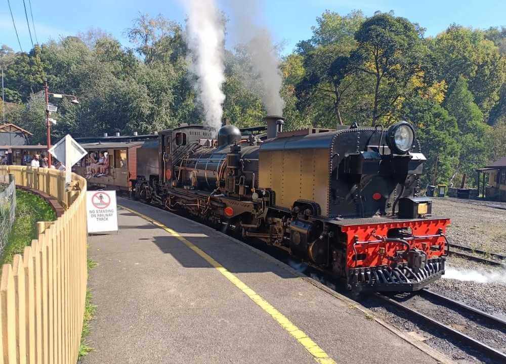A vintage steam locomotive at Puffing Billy Railway station in Melbourne, with white smoke billowing from its chimney. The black and brass engine stands on the tracks beside a wooden platform where passengers, including children, sit in open-sided carriages. The station is surrounded by lush green trees and a rustic picket fence, capturing the charm of this historic railway experience.