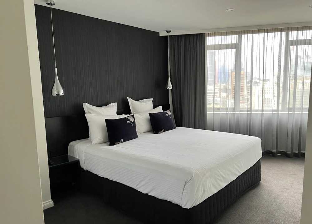 A sleek and modern bedroom at Quay West Suites Melbourne, featuring a large king-sized bed with crisp white linens, dark accent pillows, and a black feature wall. The room is softly lit with hanging pendant lights and offers a panoramic city view through large windows covered with sheer curtains.