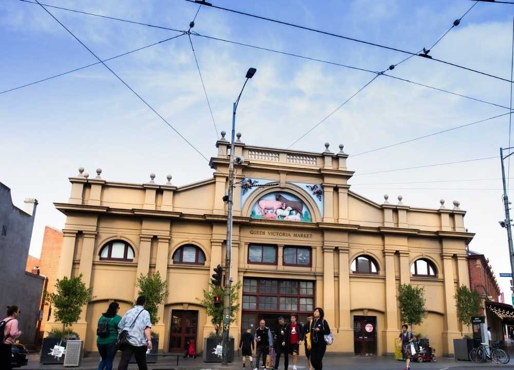 The historic Queen Victoria Market building in Melbourne, featuring its cream-coloured façade with arched windows, classical columns, and a decorative mural above the main entrance. Several people are seen walking in front of the market under a web of tram wires, capturing the lively and bustling atmosphere of this iconic shopping destination.