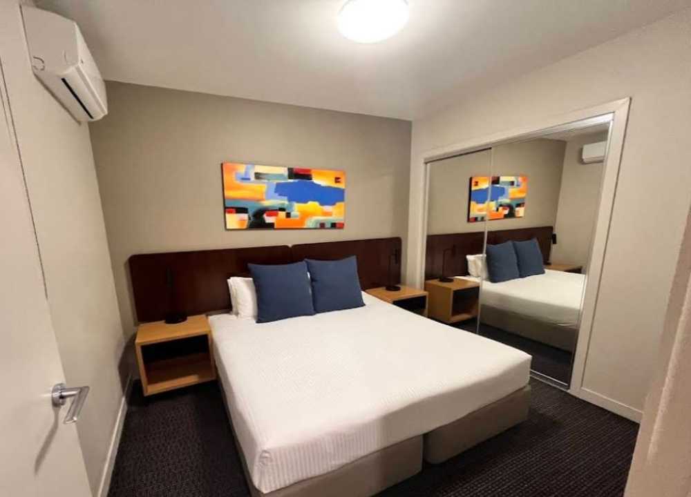 A compact and modern bedroom at Quest Docklands Apartment Hotel in Melbourne, featuring a neatly made double bed with white linens and two navy blue accent pillows. The room includes wooden nightstands with built-in lighting, a mirrored wardrobe, and a vibrant abstract artwork above the headboard, creating a clean and functional space for guests.