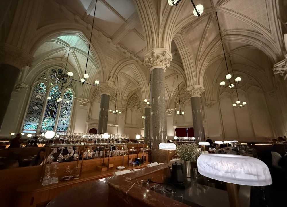 Grand interior of Reine & La Rue restaurant in Melbourne, showcasing a breathtaking neo-gothic setting with soaring vaulted ceilings, marble columns, stained glass windows, and ambient globe lighting over elegant dining booths and a central bar.