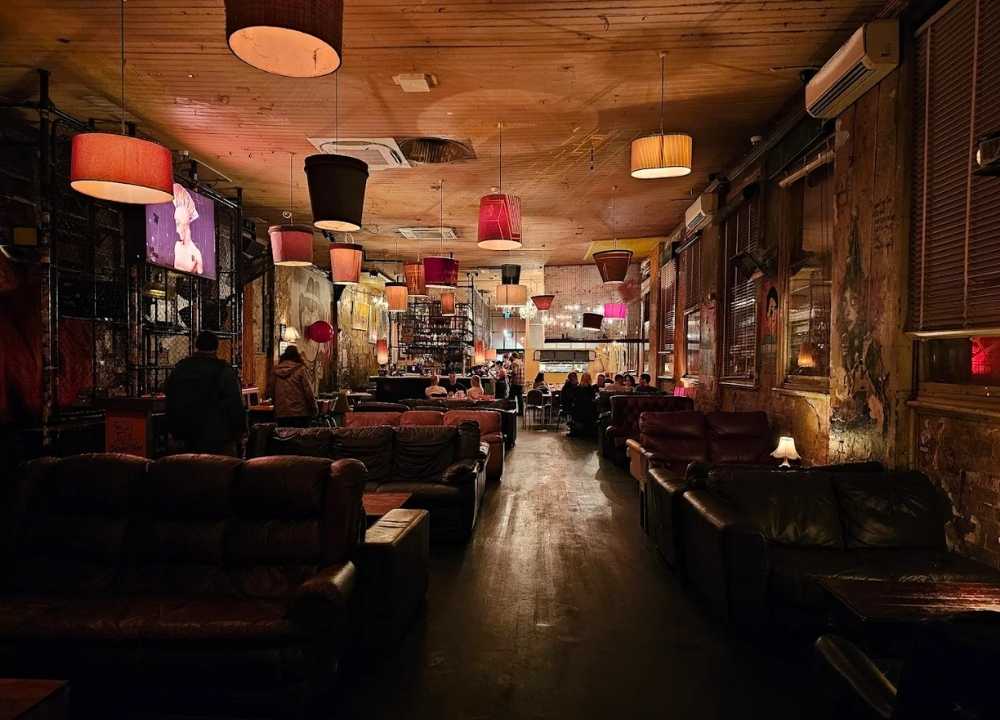 Interior of Revolver Upstairs (Revs), a well-known nightclub and lounge in Melbourne, featuring dim lighting, mismatched hanging lampshades, and worn leather couches lining the walls. The rustic décor, graffiti-covered walls, and eclectic furnishings create a laid-back, alternative vibe. Patrons are seen relaxing and socialising in the cosy, bohemian atmosphere.