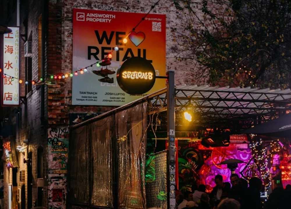 Nighttime view of Section 8, an open-air bar and club in Melbourne, located in a graffiti-covered laneway adorned with string lights and neon signs. The venue features a vibrant, urban vibe with colourful wall art, a semi-covered seating area, and a lively crowd enjoying music and drinks under glowing lights. A round sign above the entrance reads "Section 8."