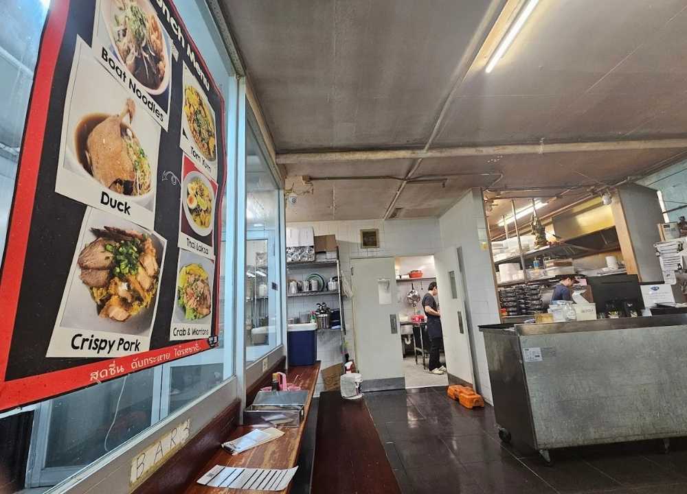 Interior of Soi 38, a hidden Thai restaurant in Melbourne, featuring a simple, industrial-style setup with exposed ceilings and a concrete floor. A vibrant lunch menu poster showcasing dishes like duck, crispy pork, and tom yum is displayed on the wall, while staff can be seen preparing meals in the open kitchen area. The casual, street-food-inspired vibe adds to its authentic dining experience.