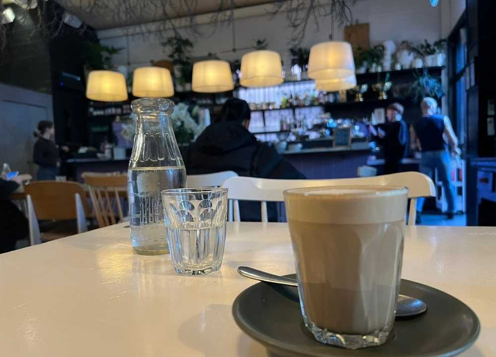 A cosy indoor scene at South of Johnston café in Melbourne, featuring a close-up of a latte served in a glass on a dark saucer with a spoon. A bottle and glass of water sit on the table, while warm pendant lights hang above a rustic interior filled with plants and shelves. Patrons can be seen chatting and ordering at the counter in the background, adding to the relaxed, welcoming vibe.