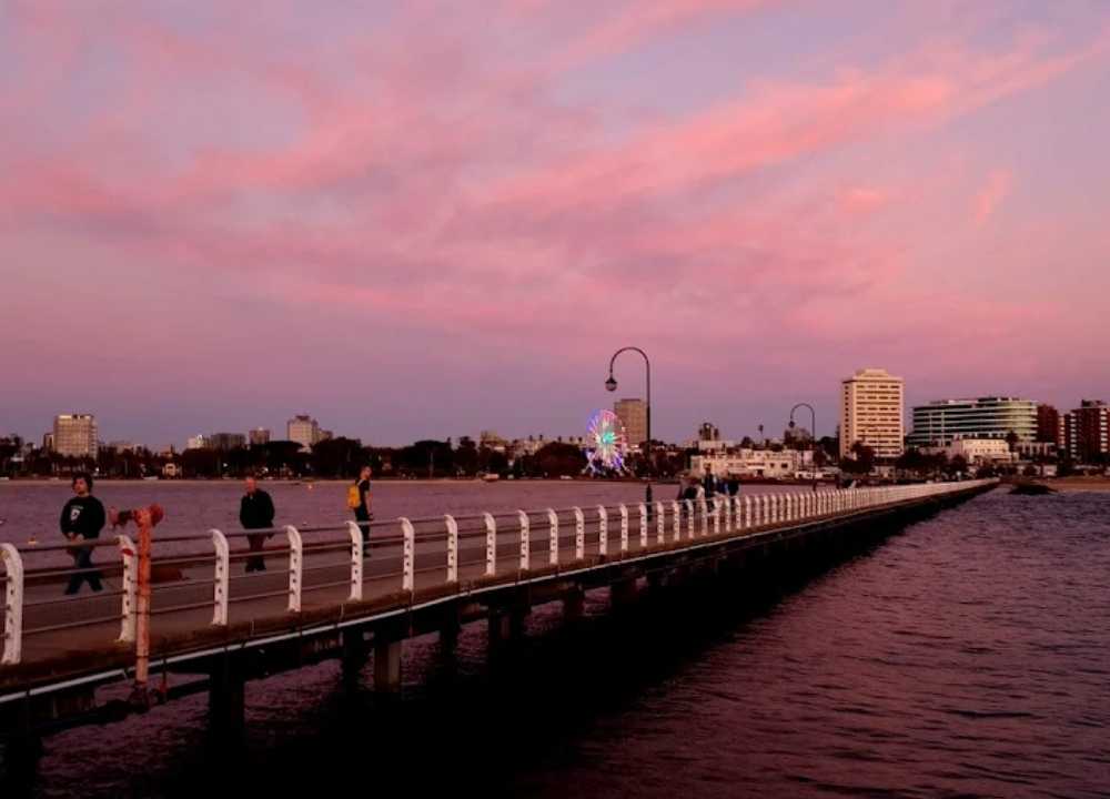 A scenic view of St Kilda Pier in Melbourne at sunset, with people strolling along the wooden boardwalk that stretches over the water. The sky is painted in soft pink and purple hues, reflecting on the calm bay. In the background, the city skyline, a glowing Ferris wheel, and waterfront buildings create a vibrant and relaxing coastal atmosphere.