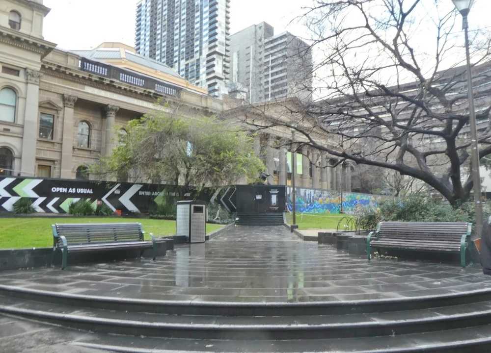 A rainy day outside the State Library of Victoria in Melbourne, showing the historic sandstone façade partially obscured by construction signage that reads "Open As Usual." The foreground features wet stone steps, park benches, and a leafless tree arching over the scene, with modern high-rise buildings towering in the background.