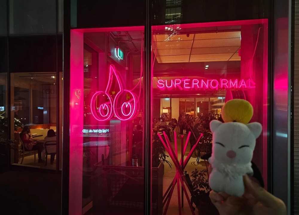 Exterior of Supernormal restaurant in Melbourne, featuring a bright pink neon sign with stylised cherries and the word "SUPERNORMAL" glowing in the window. A cute plush toy with a yellow pompom is held in the foreground, while diners are visible inside the warmly lit, modern interior.