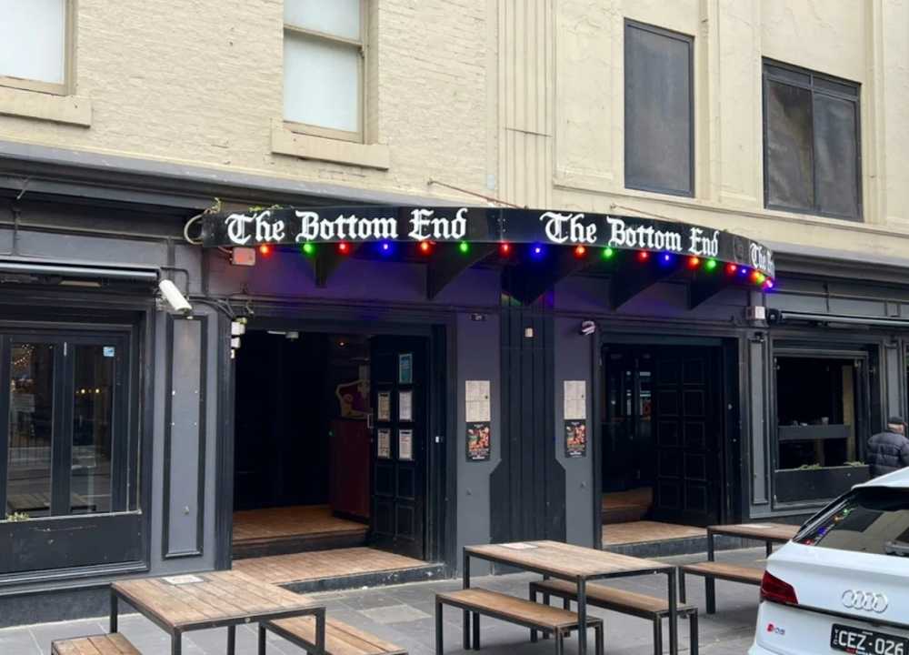 Street view of The Bottom End, a popular nightclub and bar in Melbourne. The entrance features a dark façade with a curved black awning displaying the venue’s name in gothic-style lettering, accented by colourful string lights. Wooden outdoor tables are set up on the sidewalk, and a white car is parked nearby, contributing to the casual urban vibe.