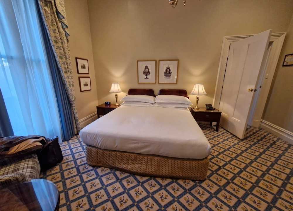 A charming vintage-style bedroom at The Hotel Windsor in Melbourne, featuring a neatly made bed with white linens and a patterned skirt. The room is decorated with classic wooden furniture, elegant table lamps, and framed artwork above the bed. The patterned carpet and floor-length curtains add to the room's historic and luxurious ambiance.
