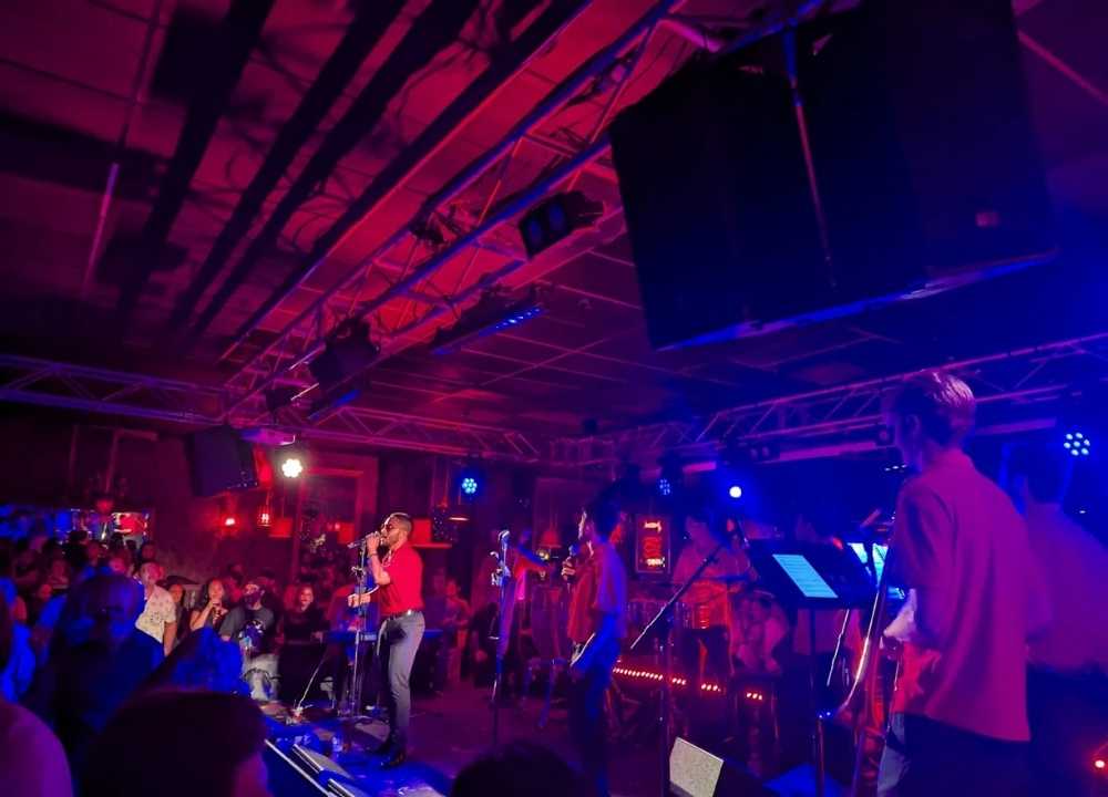 A vibrant live performance at The Night Cat, a popular music venue and nightclub in Melbourne. The stage is lit with dynamic red and blue lighting as a singer in a red shirt engages the crowd, backed by a band. The audience fills the space, creating an energetic and immersive atmosphere known for eclectic music and nightlife.