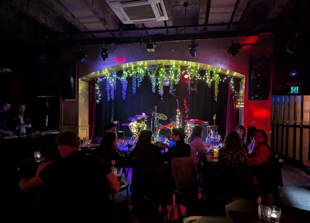 A cosy and dimly lit interior of The Toff, a popular nightclub and live music venue in Melbourne. Guests sit at candlelit tables facing a small stage adorned with hanging vines, fairy lights, and musical instruments. The atmosphere is intimate and vibrant, with red and purple lighting casting a warm glow across the room as DJs prepare in the corner.