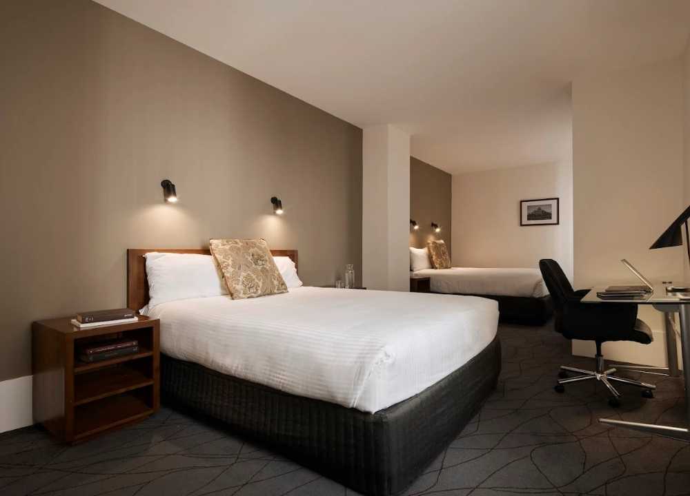 A modern and spacious guest room at The Victoria Hotel in Melbourne, featuring two neatly made queen beds with white linens, decorative pillows, and wall-mounted reading lights. The room includes a wooden nightstand, a work desk with a lamp and office chair, and a neutral-toned decor that creates a warm and inviting atmosphere for travellers.