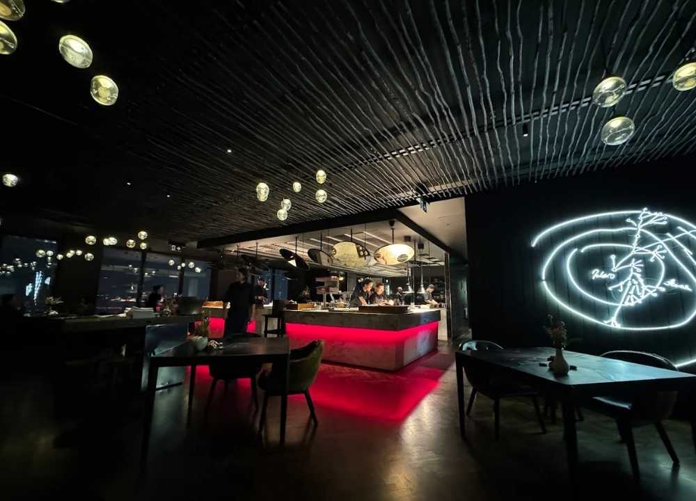 Elegant interior of Vue de Monde restaurant in Melbourne, showcasing a dramatic open kitchen with a glowing red-lit counter, modern decor, and ambient ceiling lights. A neon wall art piece adds a contemporary touch to the dimly lit, upscale dining atmosphere.